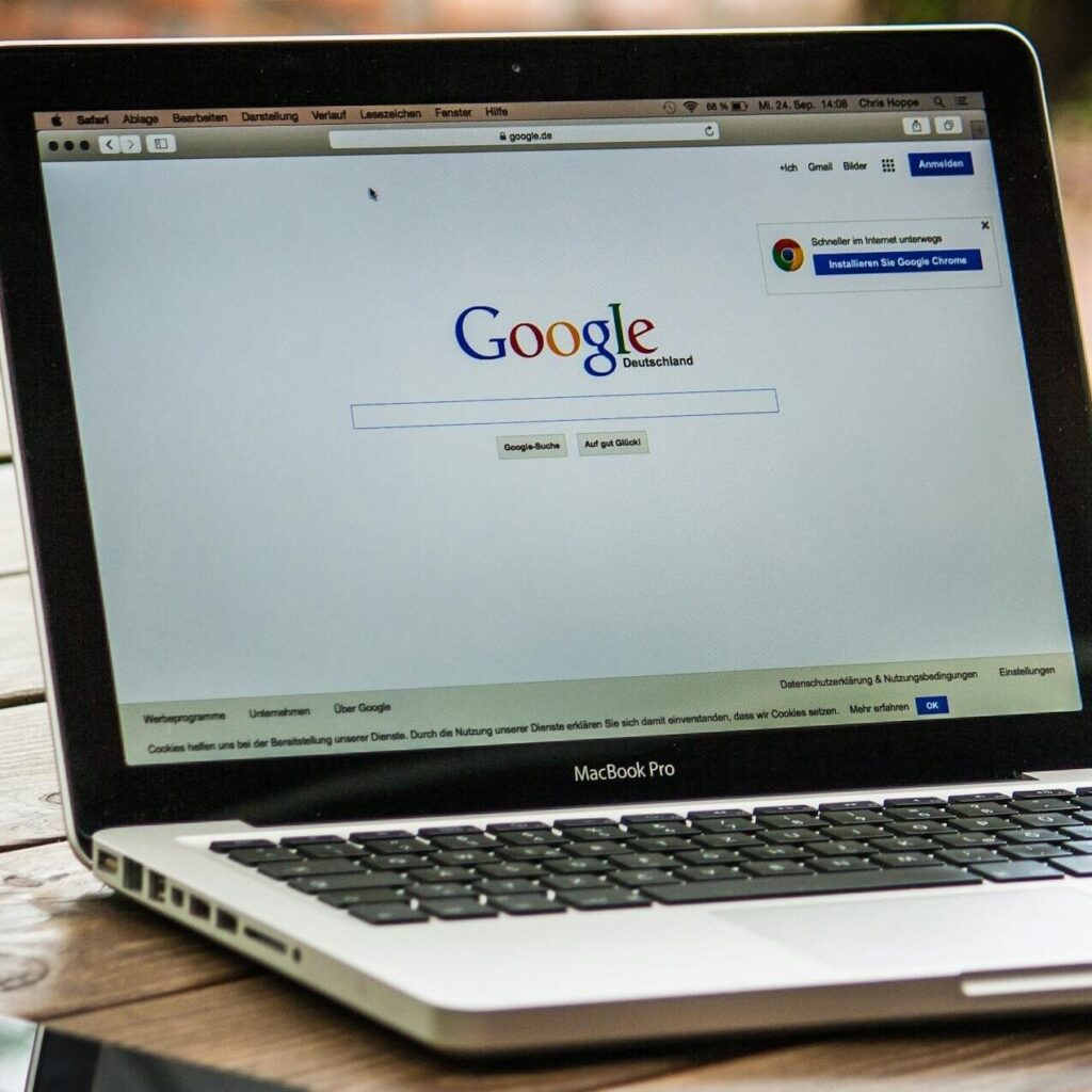 A MacBook Pro displaying Google Search on a wooden table outdoors, next to a smartphone.