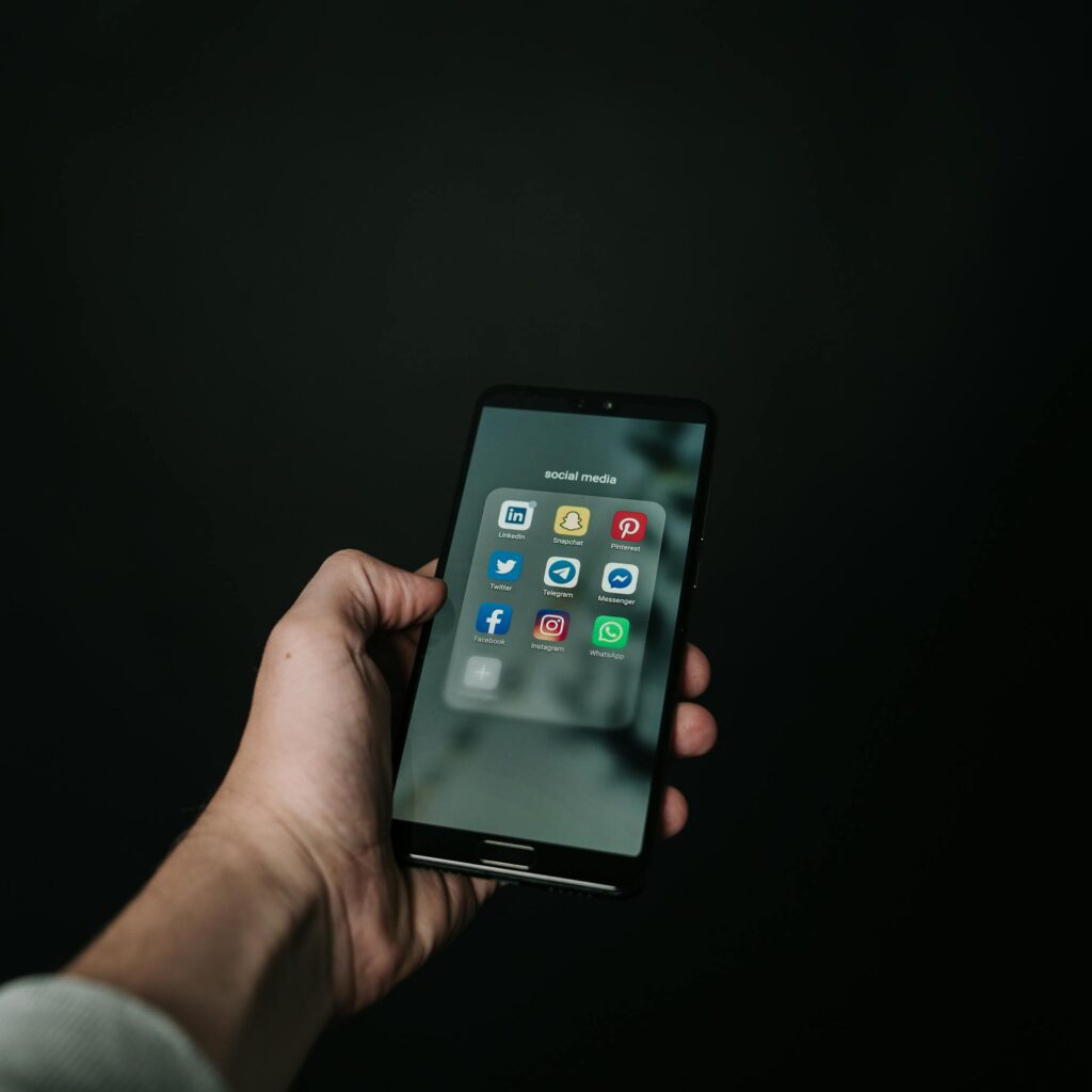 Close-up of a hand holding a smartphone displaying various social media app icons on a dark background.